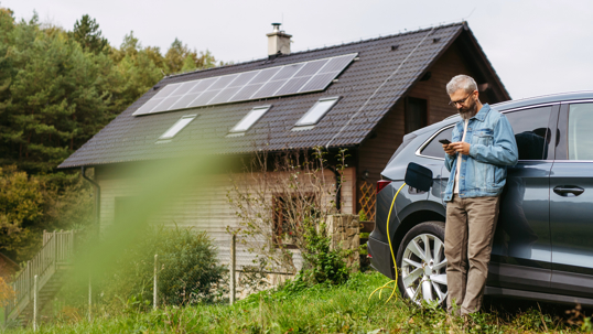 Man charging electric car in front of his house with solar panel system on roof behind him.
